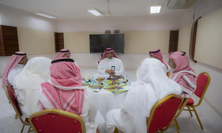 يجتمع سبعة رجال الحاضرين الغترة أو الشماغ السعودي مع العقال والبشتاء السعودي حول طاولة في جامعة القصيم، يناقشون مستندات وكتيبات. تتوزع على طاولة الضيافة وقوارير المياه، حتى تنضج شاشة كبيرة في تُبرز الاحتفاء بمناسبة اليوم العالمي للمعلم وكريم المتقدمين والمعلمات.