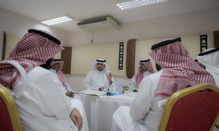 يجتمع مجموعة من الرجال مرتدين البشت السعودي والغترة أو الشماغ والعقال حول طاولة في جامعة القصيم، حيث يشاركون في نقاش علمي ضمن ورش العمل. ويبرز في تصفيته وهو يتحاور بإيماءات محددة، بينما يبرز على طاولات العمل وأكواب ضي بعض المأكولات المخصصة للحضور.