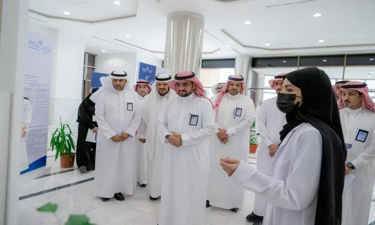 يستمع جمع من الرجال مرتدين الغترة أو الشماغ والعقال والبشت السعودي، باهتمام الكبار إلى امرأة ترتدي ملابس نسائية كما كمامة صحية أثناء تقديمها في الملتقى البحثي العلمي الذي نظمته كلي طبياً عرض في جامعة القصيم، وذلك في مبنى حديث تميز بجدرانه الأبيض ونباتات داخله.