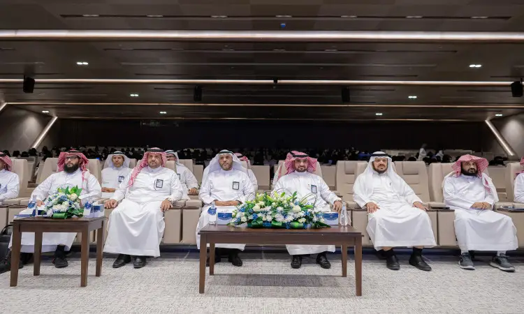 يجلسون مجموعة من الرجال مرتدين الغترة أو الشماغ والعقال والبشت السعودي في الصف الأمامي بإثنتين من القاعات الكبرى في جامعة القصيم، وذلك من خلال حضورهم الملتقى البحثي العلمي. وتزين الطاولات أمامهم زهور وقوارير، بينما يظهر الحضور في الجلوس.