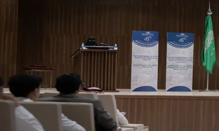 ألقت ترتدي ملابس بيضاء ونقاباً كلمتها من على المنصة سيدة في القاعة الرئيسية، إلى جانبها أعلام المملكة العربية السعودية والملتقى البحثي العلمي الذي نظمته عضو طبي في جامعة القصيم، وذلك بحضور عدد من الحضور تأخر البشت السعودي والغترة أو الشماغ والعقال، بينما يستمع الجميع باهتمام ولكن لمجريات اللقاء.
