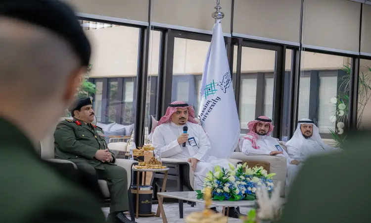 تفضل النص المعادي صياغته كما طلبت: "يتحدث رجل يرتدي البشت السعودي والترة والعقال في مكبر الصوت بينما يجلس بجانب العام رئيس جامعة القصيم والمدير للسجون، وذلك خلال اجتماع داخلي يُعنى بتمكين خريجين، جزئي في الخلفية علم المملكة وبعض الزهور.