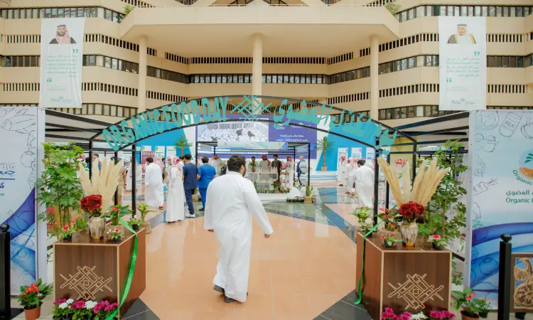 Visitors stroll through an exhibition hall decorated with banners, plants, and displays highlighting the benefits of priority foods, where a large number of attendees, dressed in a tunic or shemagh and aqal, in addition to the Saudi bisht, gather under high ceilings, distinctive architectural details, and an official public passion that has never accompanied the University of Qassim.