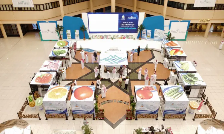 An overhead view of an indoor display featuring pavilions with insurance fruit responsibility, as part of the Promex event in Qassim. Many of the visitors in attendance are wearing the Ghutra or Shemagh and Egal, as well as the Saudi Bisht, as they wander around the spacious, brightly-lit space. It also features a large projection screen on the main stage, highlighting food ingredients and their importance in having a healthy life.