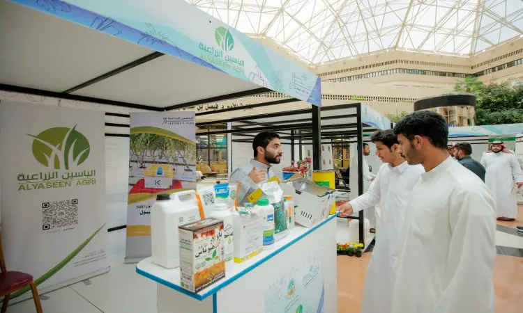 Inside a modern, glassed-in hall, three men dressed in traditional Saudi attire consisting of a ghutra or Saudi shemagh, an aqal and a Saudi bisht, walk past a representative at the produce pavilion showcasing the benefits of organic food. They are characterized by a variety of different products.