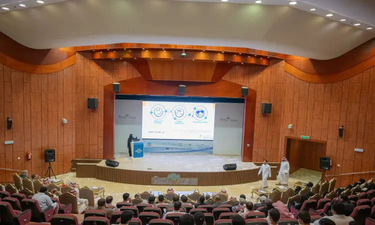 In a large lecture hall in Qassim, a large number of participants are seated in front of a platform on which the lecturer gives a clear presentation on the harms of foodstuffs. A number of people are wearing the Saudi Ghutra or Shemagh with Eqal, as well as the official Saudi Bisht.