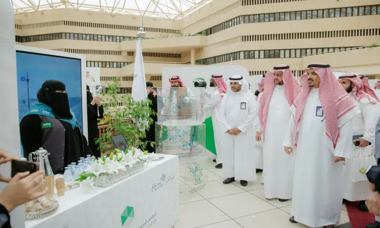 A Miami group of men in traditional Saudi attire, including the ghutra or shemagh, headband, and Saudi bisht, along with a woman wearing a jacket with a turquoise black waistcoat, at a dedicated stand inside the exhibition held on the occasion of the "Fifth Forum of Nazaha Clubs" at the university. The platform is surrounded by grass, bottles and informational materials about the exhibition.