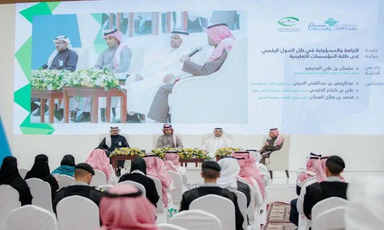 The men, dressed in the Saudi Arabian ghutra or shemagh, aqal and bisht, sit on the stage during the fourth Kitchen Forum at Qassim University, with the auditorium facing them. Behind them on a large screen is a broadcast of their image along with Arabic texts and the logos of Nazaha clubs and a number of Saudi associations.