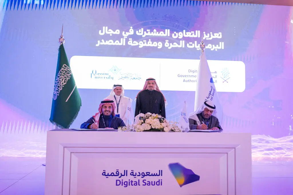 Men, two standing, participate in a signing ceremony in front of Saudi Arabian flags and a screen with Arabic text, marking the beginning of a digital partnership between Qassim University and the Digital Government Authority. The participants are dressed in the official Saudi uniform of the ghattra or shemagh with the Saudi aqal and bisht, lending a sense of official responsibility to follow the prestige of the university and the participants.