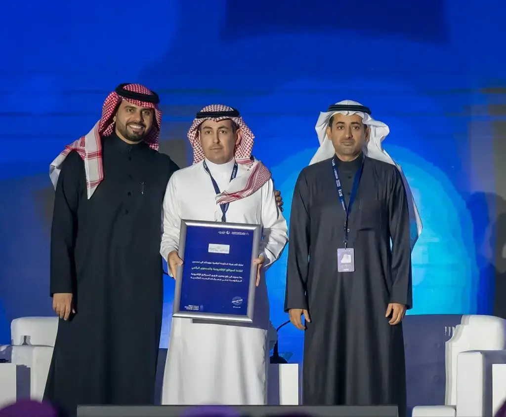 Three men dressed in a Saudi Arabian ghutra or shemagh with an aqal, and one wearing a Saudi bisht, are on stage celebrating an outstanding achievement in the field of education. The age of the man in the middle of them, who received a mediocre grade, as he smiles for the camera in full view of a backdrop lit for children in blue, in the middle of the night in Qassim.