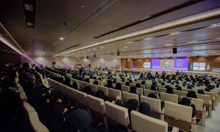 قاعة مؤتمرات واسعة في جامعة القصيم تغص بالحضور، حيث يجلس العديد من الرجال مرتدين الشماغوقال والبشت السعودي، إلى جانب النساء ارتدين الأعباء السوداء والنقاب. جميعهم متجهون نحو المنصة التي تتوسطها الألوان للعرض ولافتات تحت أضواء سقفية ساطعة، وذلك ضمن مؤسسة رسمية بإنجازات الطلاب في يوم الطالب.