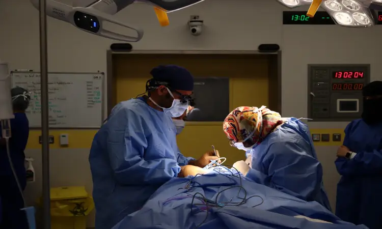 Two surgeons dressed in blue surgical scrubs with a Saudi shemagh and a Saudi bisht are gathered in the specialized operating room of Medical City University, performing the delicate surgical tasks for the patient, while a specialized medical team and modern medical equipment support them to provide the necessary vital services in the background.
