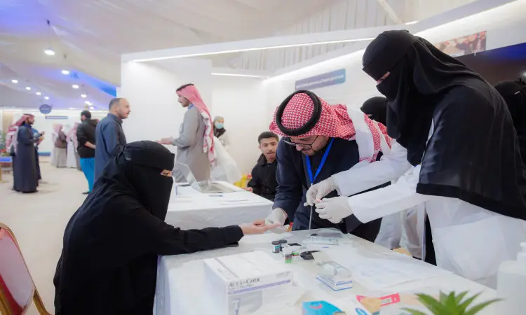 تجلس ترتدي ملابس بيضاء ونقابية على طاولة في جناح طبي تابعة لمركز دخنة، حيث يقوم أحد العاملين في الحماية الصحية بالإدانة بفحص دمها ضمن فعاليات التوعية والتثقيف. في الخلفية عدد من الحضور الحضور البشت السعودي، والشماغ السعودي أو الغترة السعودية مع العقال، في صورة الاحترام الطابع الرسمي والمتمسكة بأصالة الملبس الوطني داخل جامعة القصيم.