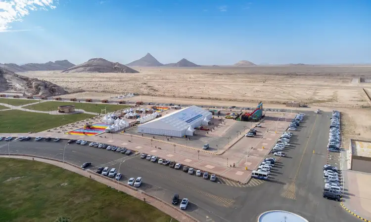 Under a clear blue sky in the middle of a desert area with rocky hills, a large tent has been erected with many creative people as part of Qassim University's awareness and education campaign. Dressed in the Saudi Arabian ghutra or shemagh, aqal, and pashtina, participants gather to welcome women to the Kingdom's authentic heritage, while cars are lined up near the event site amidst the view and tranquil surroundings.