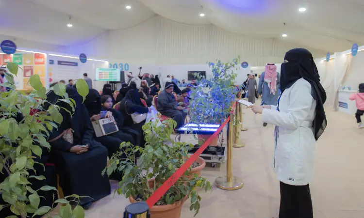 A Palestinian woman wearing a medical coat and a union coat stands among the attendees of the health awareness and education campaign organized by Qassim University at the Dukhna Center. A number of informational corners are visible in the background, while some attendees are wearing the Saudi shemagh, aqal and bisht, respecting the official faith of the event, which focuses on promoting health and cultural awareness in the community.
