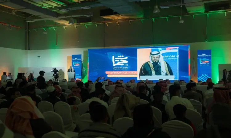 يجلس جمع غفير من الحضور في قاعة المؤتمرات، ويواجه المنصة التي يظهر عليها صاحب السمو أمير منطقة القصيم عبر الشاشة مرتديًا الغترة السعودية والعقال والبشت السعودي. سموه اختاره خلال مهرجان الجامعة، بينما تضيء أرجاء القاعة أنوار خضراء وحمراء وتلتقط صور الكاميرات لحظات الاحتفاء بتراث الشعوب.