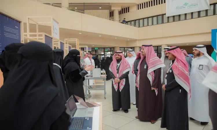 يجتمع مجموعة من الرجال في زيهم السعودي التقليدي، حيث يرتدي الرجال الغترة أو الشماغ والعقال والبشت السعودي، حول طاولة العرض في معرض المبدعين داخل مبنى حديث ومضاء جيدًا. تقوم إحدى السيدات بتقديم المعلومات، بينما يستمع للآفات باهتمام، ومن بينهم معالي رئيس جامعة القصيم.