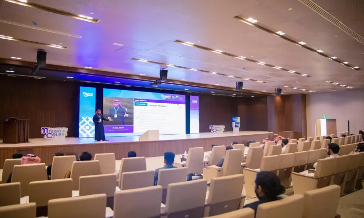 He takes the stage at the Modern Hall in Qassim, dressed in the Saudi bisht and aqal, while large screens are projected behind him around a giant stage. Medical students in Saudi uniforms and traditional hangers in rows of cream-colored chairs, maintaining proper spacing until the women's costumes are distinguished.
