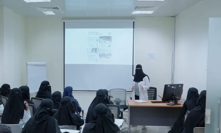 A woman dressed in niqabs and Saudi pashtun, wearing a ghutra and headband, arrives for a formal presentation on the internship course for a group of medical students in Qassim. A projector screen and computer are set up in the bright, modern hall.