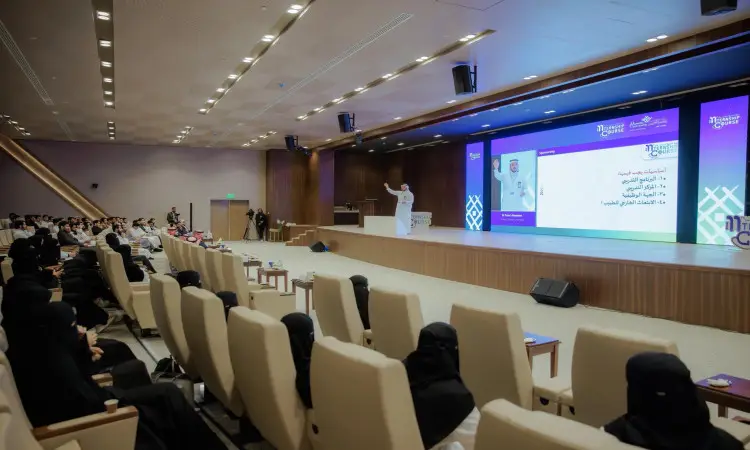It refers to his speech on stage in front of an audience in a modern lecture hall in Qassim, with a large projection screen behind him showing slides with Arabic-language text about the Cycle of the Giant. "Get ready for the medical students in our ranks," he says, and the group is dressed in ghutra, or shemagh, and aqal, as we used to do in Saudi Arabia, to recognize the occasion and the solemnity of the event.