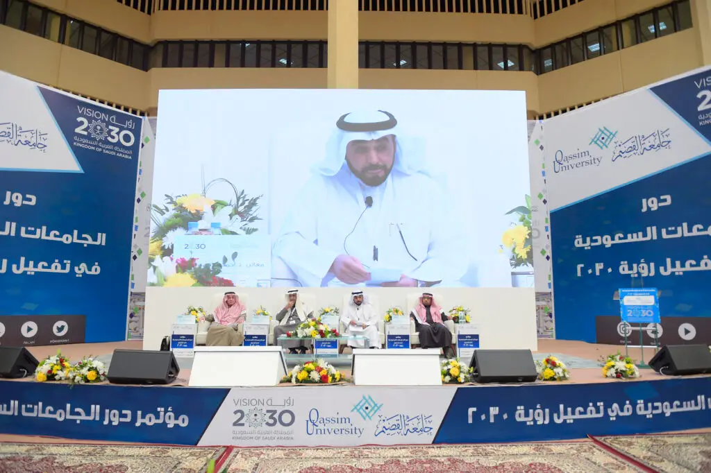 جلسة أربعة رجال الغترة أو الشماغ السعودي والعقال والبشتاء السعودي على المنصة خلال مؤتمر في جامعة القصيم حول التعليم الإلكتروني. وقد ظهر خلفهم شاشة كبيرة تُبرز البارزين، بينما تزين القاعة لافتات تحمل شعاري رؤية 2030 باللغتين العربية والإنجليزية.