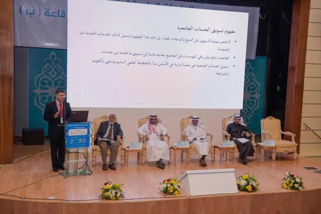 كل خمسة رجال لبس الغترة والعقال والبشت السعودي على منصة العرض أمام شاشة كبيرة تعرض نصاً عربياً حول رؤية 2030، وذلك من خلال مؤتمرات مؤتمر في جامعة القصيم. بوضوح أحدهم من خلف المنصة، حيث جلسوا وبحوزتهم مستندات رسمية. وتزين باقات الزهور، لتضفي جواً من الفخامة وتفضل بمكانة السعودية.