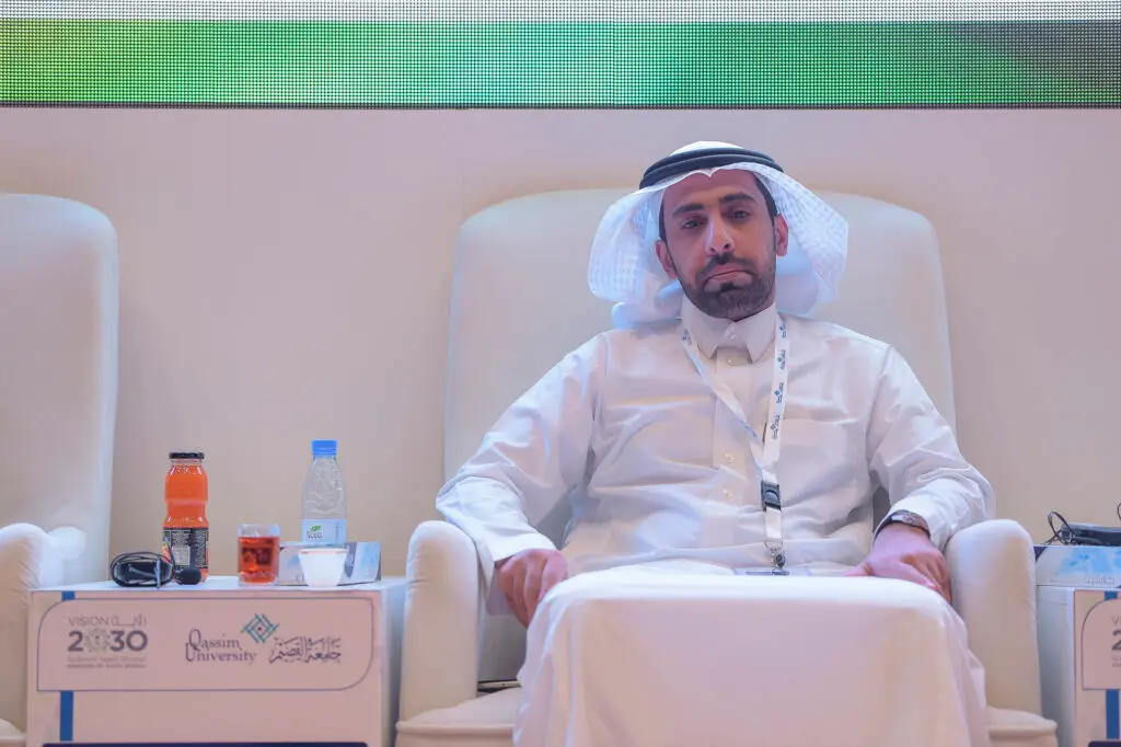 يجلس رجل مرتدياً كرسي الغترة والعقال والبشت السعودي على الأبيض، للتأكد من جواره مشروبات ولوحة اسمية، وذلك من خلال فعالية لجامعة القصيم تبرز شعار رؤية 2030 وتركز على تحسين البيئة التعليمية ومخرجات التعليم العالي.