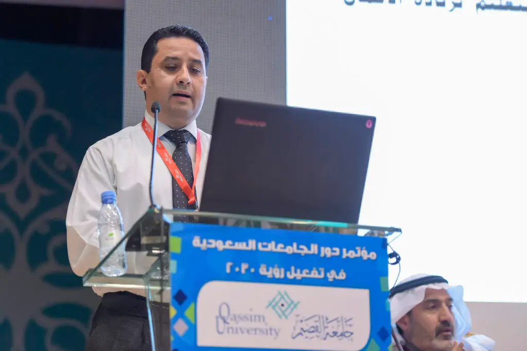 A man wearing a white shirt and tie is at the conference podium, in front of his laptop, reviewing the professional education themes of the Qassim conference. A man sits next to another man wearing a Saudi Arabian ghutra, headband and bisht, while a large screen behind them shows texts related to the event.