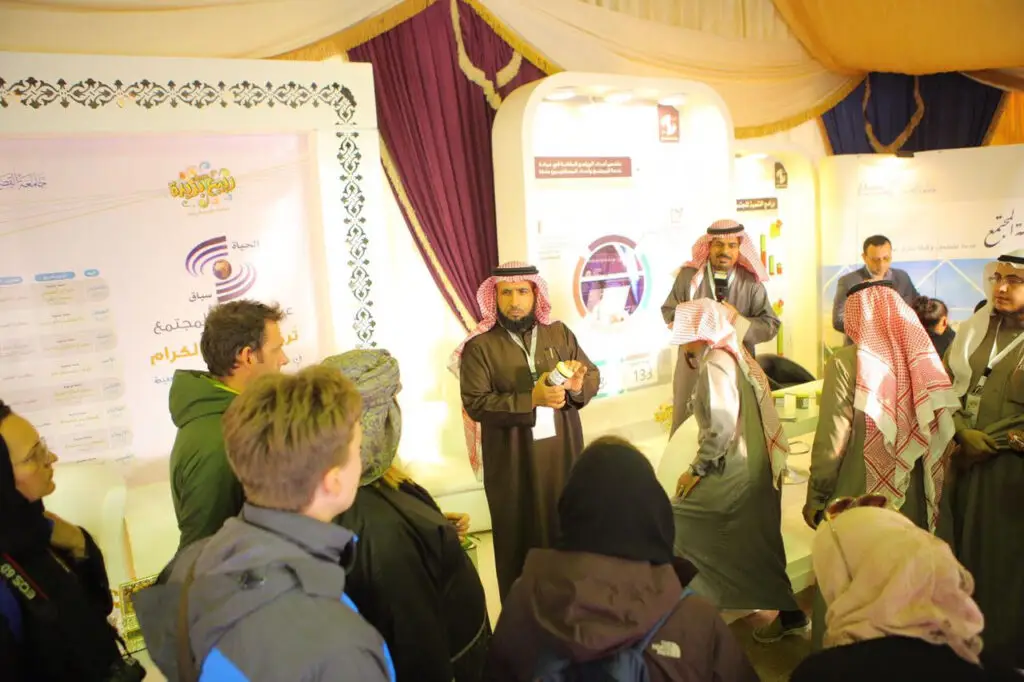 A group of men dressed in formal Saudi attire, including the Ghutrah or Shemagh, Eqal and Saudi Bisht, interact with the winter clothing brigades in a brightly lit exhibition stand for the health education campaign. The Qassim College of Fisheries' logos are featured alongside informational and educational displays in Arabic.