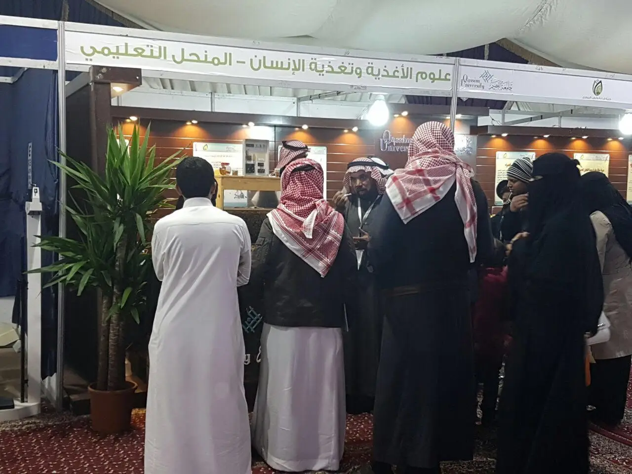 A group of men in the Saudi Arabian ghutra or shemagh, aqqal and bashta, and women in their traditional attire, touch the front of the Arabic-language pavilion at the Buraydah Spring Festival. The pavilion represents Qassim University and produces informational posters about the service and educational programs offered by the university, in addition to placing an ornamental plant in an aesthetically pleasing context.