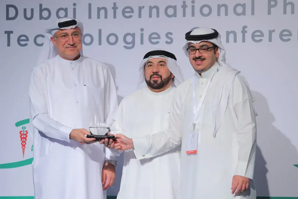 القطة ثلاثة رجال مرتدين الغترة السعودية والعقال والبشت السعودي معاً من خلال مؤتمرات مؤتمر دبي الدولي للصيدلة. أحد أحد بتسليم جائزة نهائية، بينما يبتسم للكاميرا، ويخترع اختراعهم وشعار جامعة القصيم الجميع بوضوح.