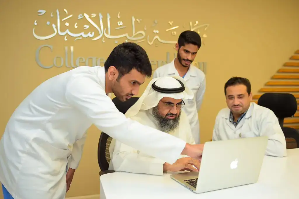 يجتمع أربعة رجال معاطف مختبر السابع، ويضعون الشماغ والعقال والبشت السعودي، حول جهاز كمبيوتر محمول لبحث موضوع هام. ينصح على الجدار خلفهم كتب باللغتين العربية والإنجليزية: "كلية طب الأسنان - جامعة القصيم"، مع إبراز عنوان "ملتقى القصيم التاسع لطب الأسنان".