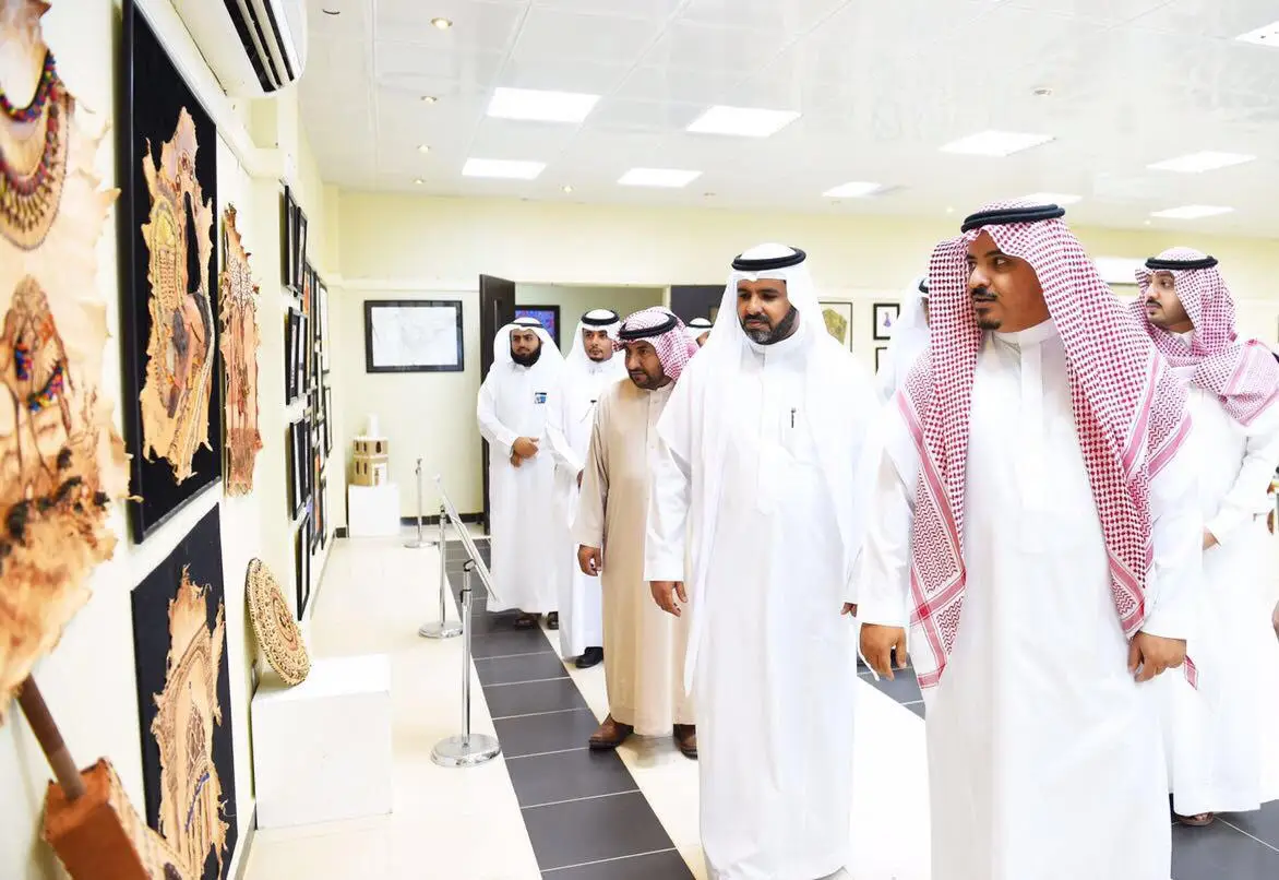 مجموعة من الرجال يرتدون الزي السعودي التقليدي - بما في ذلك الغترة أو الشماغ السعودي، والعقال، والبشت السعودي - يراقبون القطع الفنية المعروضة على جدران المعرض خلال معرض فن، الذي ينظمه كلية التصاميم والأسواق المنزلية في القصيم. يعكس هذا الواقع روح رؤية 2030 في قاعة عرض مضاءة بإتقان ومهيأة لاستقبال الزوار.