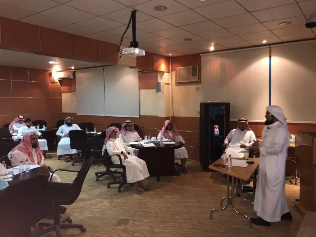 يجتمع مجموعة من الرجال مرتدين الغترة أو الشماغ السعودي مع العقال والبشت السعودي حول عدد من الطاولات في قاعة مؤتمرات في القصيم، حيث يتابعون باهتمام عرضًا يقدمه أحدين بالقرب من المنصة. تتضمن هذه اللائحة البنود الواردة في الوثيقة الأساسية خلال برنامج نصف القصيم، كما يلاحظ وجود عرض مختصر لمعلق لدعم العرض التقديمي.