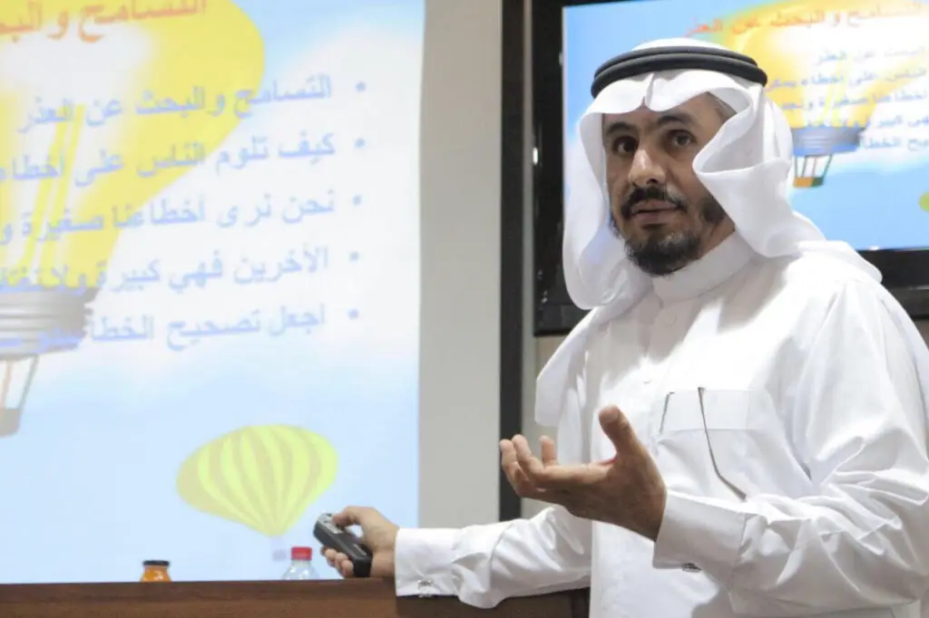 يرتدي رجل غترة سعودية وعقالاً بشتاء سعودي رسمي، ويقدم عرضًا حول درجات البكالوريوس في جامعة القصيم مستخدمًا الشمّاع للركاب. تحتوي على شريحة معروضة على نص عربي ورسومات ملونة لمنطاد الهوائي، تُبرز فعالية "صيف القصيم".
