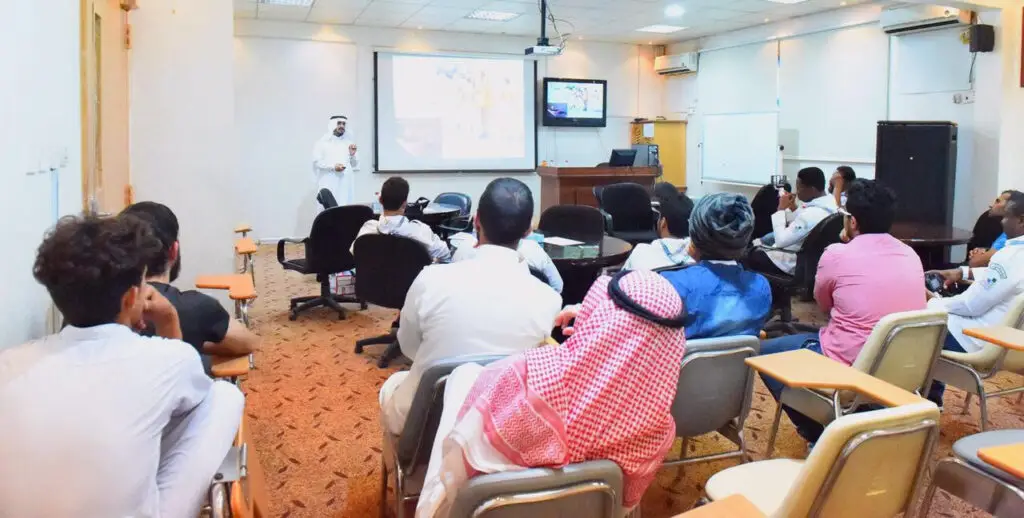 يحضر مجموعة من المشاركين في جامعة القصيم، حيث يستعدون في القاعة ويحضرون معظمهم الغترة أو الشماغ مع العقال والبشتاء السعودي، في سينعكس الحضور حيث. اتصل المحاضر بجوار شاشة عرض كبيرة للمحتوى البصري الملهم، بينما يجتمع الجميع باهتمام الكبار؛ ليجسدوا روح التعلم الصيفي المتميز في صيف القصيم.
