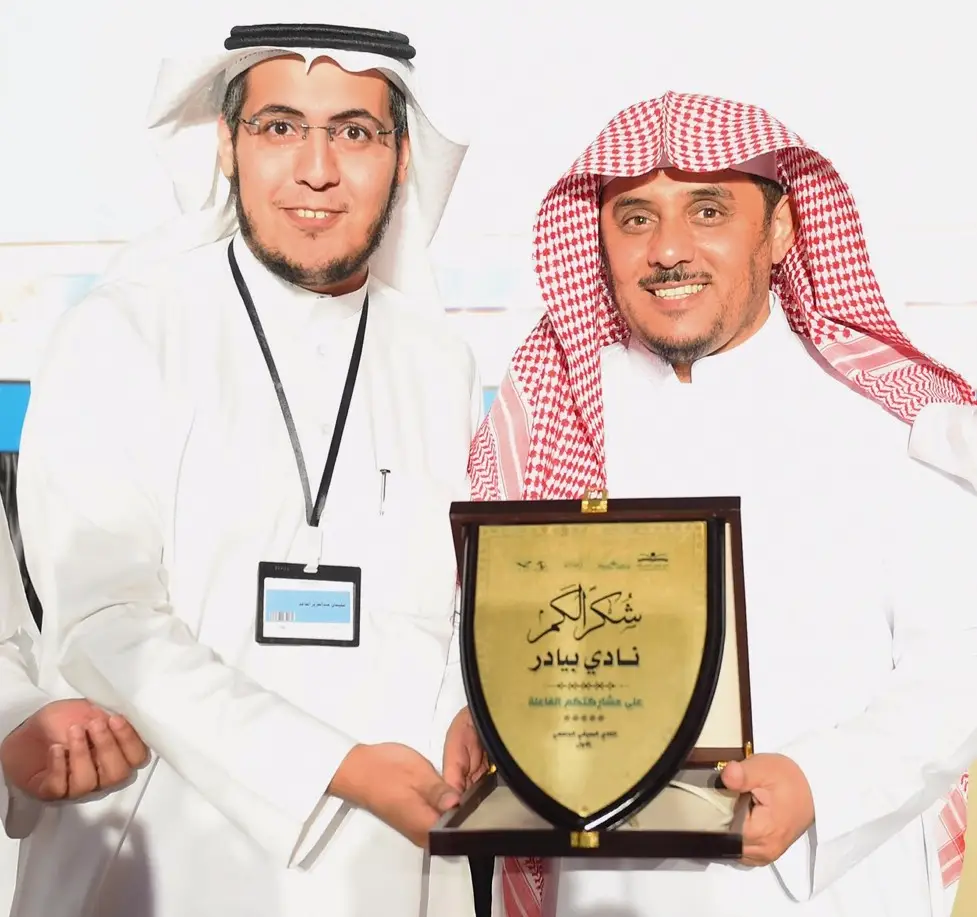 ويظهر في الرجل العباءة يرتديان اللباس السعودي التقليدي، حيث يرتدي كل منهم الشماغ والعقال، بينما يرتدي أيضاً البشت السعودي. يبتسم الرجلان أمام الكاميرا خلال إحدى المناسبات المميزة الخاصة بالنادي الصيفي" في جامعة القصيم. تركز على درعاً كتبت عليه عبارات باللغة العربية لإنجازاتهما في الجامعة، بينما يظهر البعض واضعاً ويحمل بطاقة تعريفية للسيدات، معبرين لذلك فخرهما واعتزازهما تحقق بما في نجاحات ضمن فعاليات وبرامج النادي الصيفي بالجامعة.