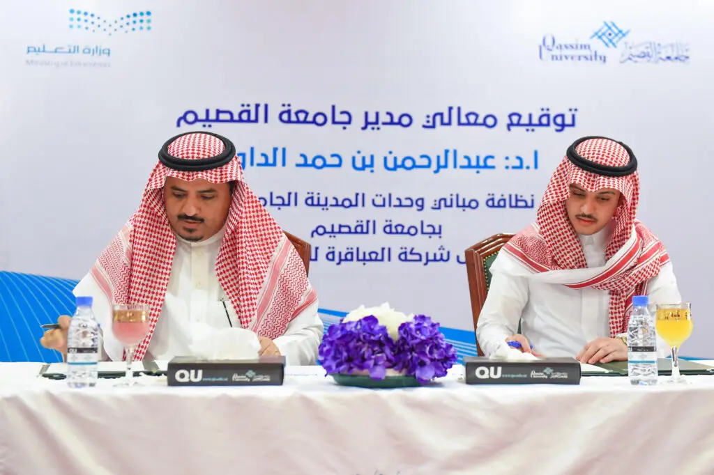 يجلس رجلان يرتدي الزي السعودي الرسمي المكون من الشماغ أو الغترة، والعقال، والبشت السعودي حول طاولة، وعليهان بتوقيع عقود تطوير جامعة القصيم. للشرب في الخلفية شعارات جامعة القصيم ووزارة التعليم، بالإضافة إلى نص عربي يُشير إلى التوقيع. كما تتواجد على جدول تنسيق مياه الشرب والميكروفونات مما يعكس الحقيقة.