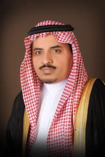 The contact man is wearing the official Saudi dress, a black Saudi bisht with gold thread, and a red and white Saudi shemagh with an aqqal, in front of a stable background. His features are marked by the active pride of the Hajj season under the patronage of the Custodian of the Two Holy Mosques.