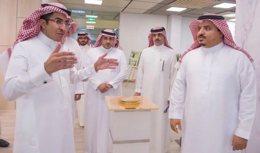 مجموعة من الرجال لبس الغترة والعقال والبشتاء السعودي الاتصالون داخل إحدى أغطية القاعات في بيئة مكتبية حديثة في القصيم، يتبادلون الحوار حول موضوع الاقتصاد. ويظهر أحد وهو يشرح وجهة نظره مستخدمًا الإيماءات بيديه، بينما تقضيت الآفات باهتمام وجدية.