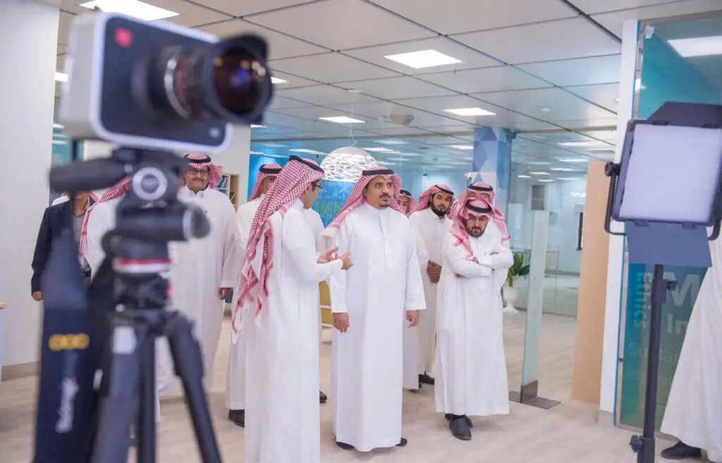 يحضر مجموعة من الرجال الحاضرين الغترة أو الشماغ والعقال والبشت السعودي، ومن بينهم الدكتور الداود، وهم يتحاورون في مكتب عصري حول موضوع الاقتصاد والدراسات العليا. خرجت في تفاصيل الكاميرا وإضاءة، ما يوحي أن هناك جولة مصورة أو تجريب توثيقه داخل جامعة القصيم.