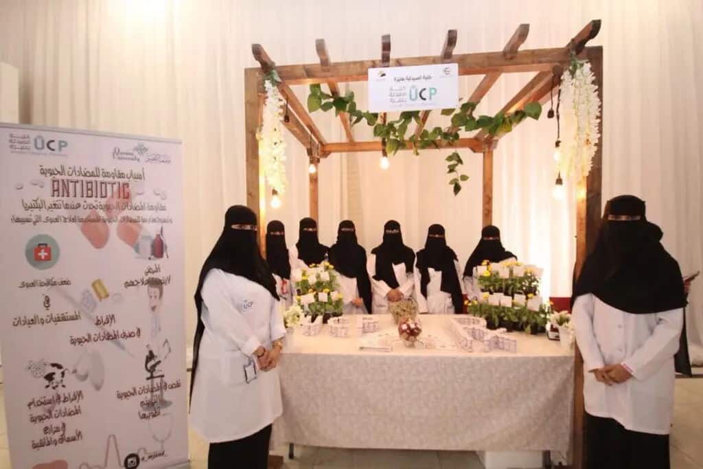 مجموعة من، يرتدون المعاطف البيضاء ويغطون وجوههن بالنقاب، يتواجدون في جناح مزدان بالزهور والنباتات. ويقفن متوشحات الأرنبة أو الشماغ والعقال والبشت السعودي بصورة لطيفة تليق بمكانتهن الأكاديمية. احترس خلفهن لوحات تعريفية حول الأنشطة الحيوية التي تحمل شعارات كلية الصيدلة بعنيزة خلال فعاليات الأسبوع العالمي للتوعية بالمضادات الحيوية، الذي ينظمه جامعة القصيم الجرايد يبرز القيم الوطنية والهوية السعودية.