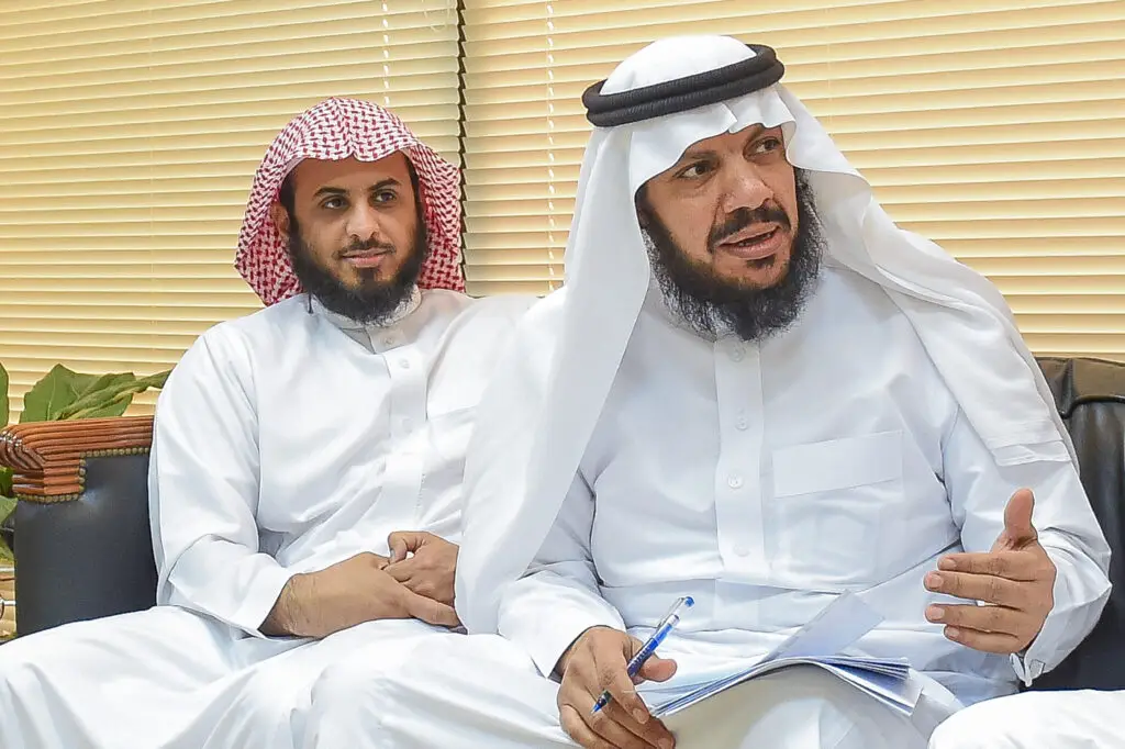 يجلس رجلان ويرتدي الغترة أو الشماغ السعودي مع العقال والبشت السعودي في مكان مغلق، حيث يركز على أوراقها وقلمًا رسميًا ويناقش سير العمل بشكل جزئي، بينما يصغى الآخرون باهتمام. تاخد في النافذة الخلفية ستائر.