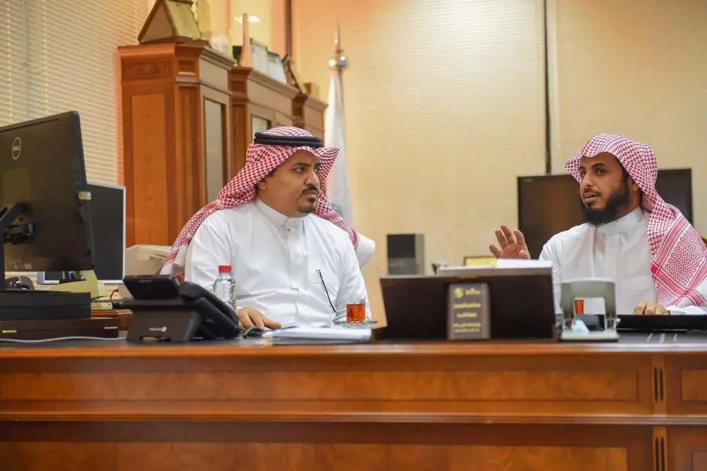ارتدى رجلان البشت السعودي، والغترة أو الشماغ مع العقال، على مكتب خشبي كبير في أحد الطلاب الرسميين في القصيم. تتوزع أمامهما أجهزة الكمبيوتر والوثائق فاجين الشاي أثناء مناقشتهما لسير العمل؛ حيث يشرح النقاط التجارية بإيماءات محددة، بينما يُنصت الآخر باهتمام وجدية.