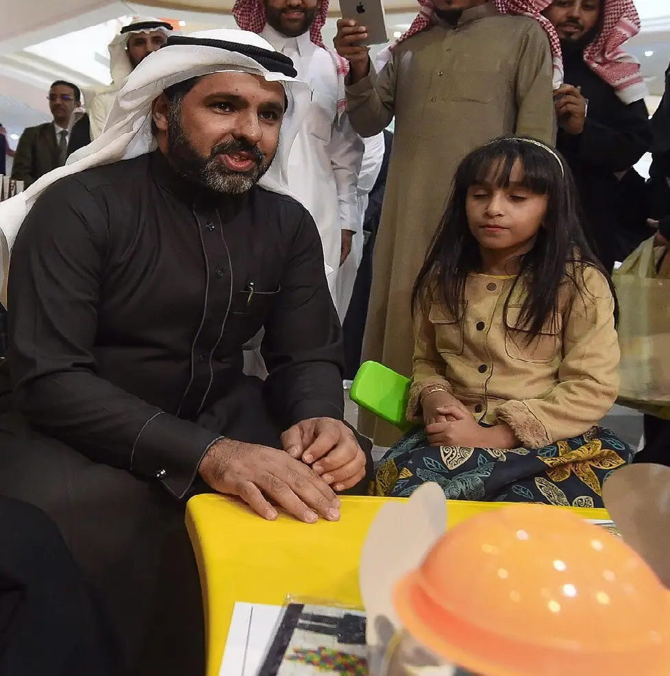 يجلس رجل يرتدي الغترة السعودية مع الشماغ والعقال والبشت السعودي بجوار فتاة صغيرة على الطاولة الصفراء، وذلك من خلال حملة توعوية في كلية التربية في القصيم، وسط حضور عدد من الناس. يبدو الرجل منتبهاً أثناء تحسين ملامح التفكير في الفتاة في هذا المكان الداخلي العام.