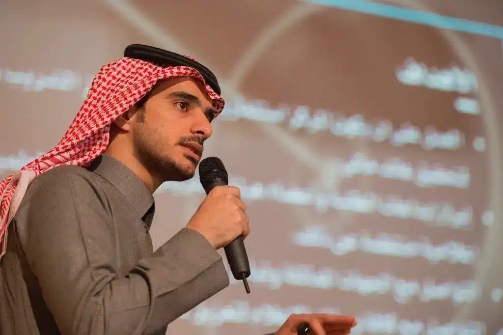 رجل يرتدي عادة غترة سعودية وعقالاً وبشت سعودي رسمي، ممسكاً اً أمام شاشة غير واضحة المعالم لتحدث نصوصاً باللغة العربية، وذلك من خلال نظمها الفعالة لمركز الإعلام والاتصال في القصيم حول تقارير النشر الإلكتروني.