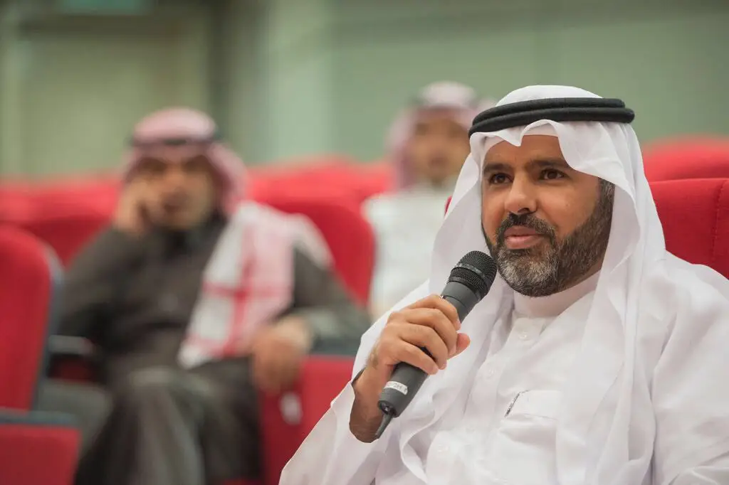 يجلس رجل ويرتدي الغترة السعودية والعقال والبشت السعودي أثناء الضغط على ميكروفون ويتحدث خلال إحدى فعاليات مركز الإعلام والاتصال في القاعة الحمراء في القصيم. ونتيجة لذلك، خارج النطاق البصري الكامل، رجلان آخر يرتديان الشماغ السعودي والعقال والبشت السعودي.
