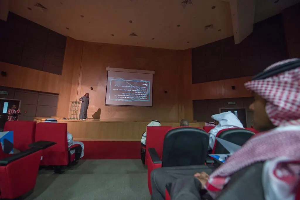 يعتلي المنصة في قاعة الاحتفالات، حيث خلفه شريحة العرض المرئي، فيما يجلس الحضور منصتين باهتمام العجوز. يبرز منهم عددٌ من الرجال مرتدين الغترة السعودية أو الشماغ مع العقال والبشت السعودي التقليدي. ولهذا السبب الرسمي، ليؤكد على أهمية إعلامية عديدة للجامعات وسياسة النشر الإلكتروني في جامعة القصيم.