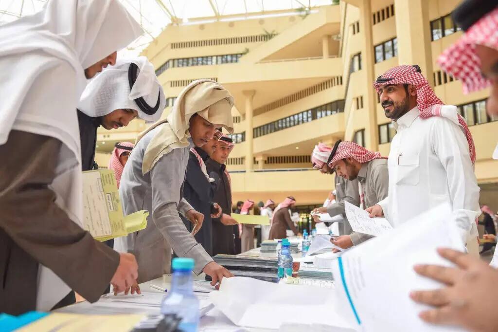 مجموعة من الرجال الحاضرين الغترة أو الشماغ والعقال، وبعضهم يلبس البشت السعودي، يتفاعلون تماما ويستعرضون وثائق التخرج على طاولة التسجيل داخل مبنى جامعي حديث وواسع في جامعة القصيم. يجب أن يتم تحديد فترات زمنية محددة للمياه وأوراقها المتعلقة بالعقوبات الجزئية للطالبات.