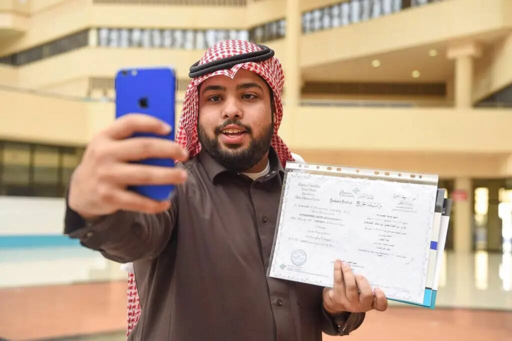 رجل يرتدي غترة سعودية وعشت رسمياً في إحدى القاعات والواسعة، ممسكًا بوثائق البراءة الحديثة، ويلتقط صورة ذاتية بهاتفه المحمول الآخر، مُحتفيًا بإنجازه كأحد طلاب جامعة القسيم.